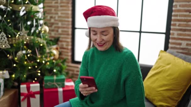 Mujer hispana madura con cabello gris usando un smartphone sentado en el sofá junto al árbol de navidad en casa - Metraje, vídeo