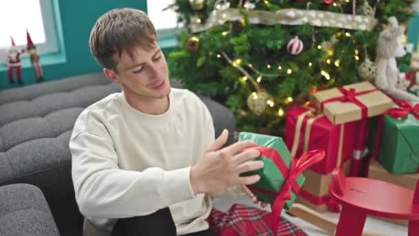 Νεαρός καυκάσιος άνδρας δίνει χριστουγεννιάτικο δώρο κάθεται στο πάτωμα στο σπίτι - Πλάνα, βίντεο