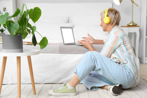 Mature woman with headphones learning English language online in bedroom - Φωτογραφία, εικόνα