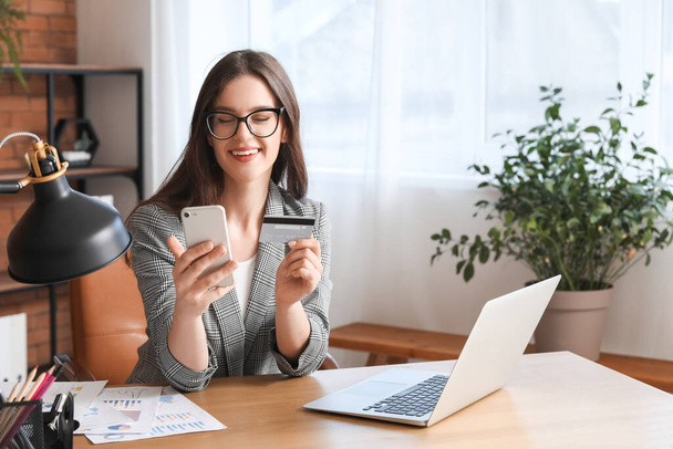 Молода бізнес-леді з кредитною карткою, використовуючи мобільний телефон за столом в офісі - Фото, зображення