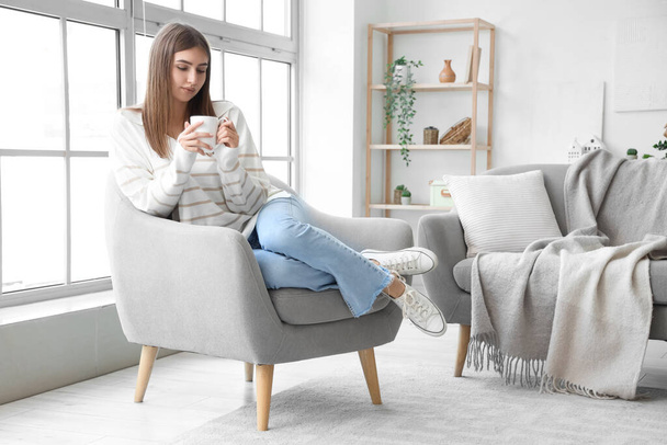 Młoda kobieta z filiżanką gorącej herbaty siedzi w fotelu w domu - Zdjęcie, obraz