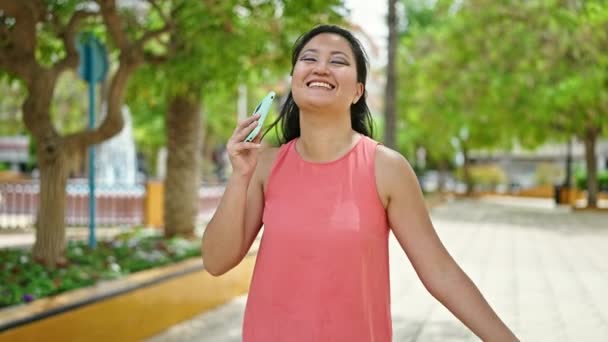 Молода китайська жінка танцює слухаючи музику на смартфоні в парку - Кадри, відео