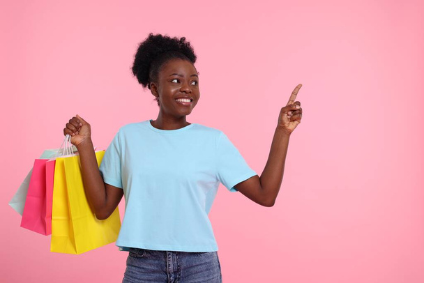 Happy young woman with shopping bags pointing at something on pink background. Space for text - Fotografie, Obrázek