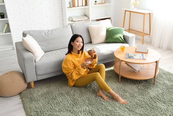 Kaunis nuori aasialainen nainen kuppi teetä istuu lattialla olohuoneessa - Valokuva, kuva