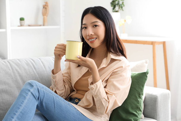 リビングルームにソファーに座っている紅茶のカップを持つ美しい若いアジアの女性 - 写真・画像