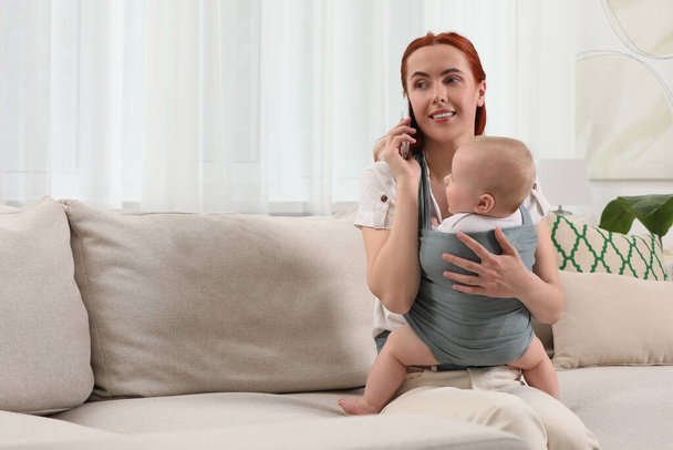Mother talking on smartphone while holding her child in sling (baby carrier) at home - Fotografie, Obrázek