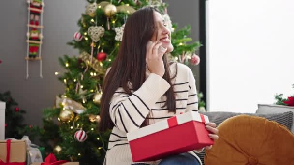 Νεαρή όμορφη ισπανόφωνη γυναίκα κρατώντας χριστουγεννιάτικο δώρο μιλώντας στο smartphone στο σπίτι - Πλάνα, βίντεο
