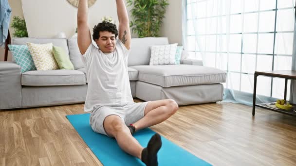 Молодой латинос, сидящий на полу, тренирует йогу, растягивая ноги дома. - Кадры, видео