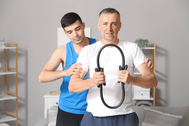 Hombre maduro entrenando con pilates círculo y terapeuta de rehabilitación en casa - Foto, Imagen
