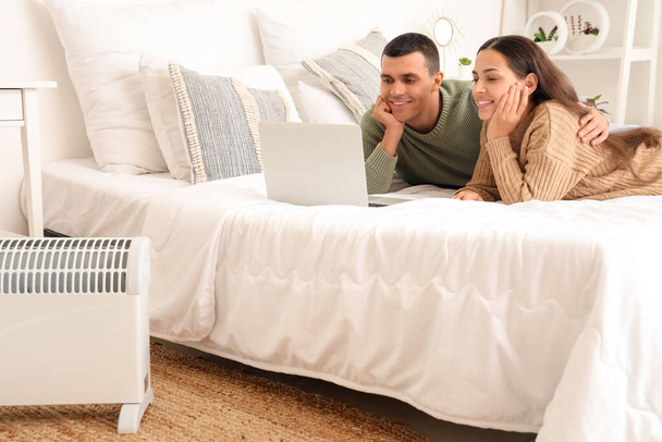 Молодая пара с ноутбуком согревается возле радиатора в спальне - Фото, изображение