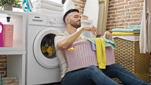 Молодий іспаномовний чоловік тримає кошик з одягом, спираючись на пральну машинку, втомився у пральні - Кадри, відео