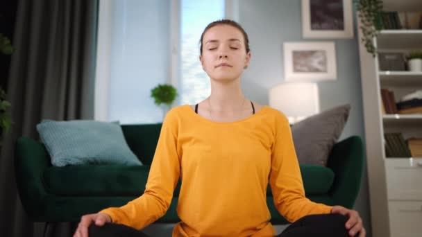 Jonge vrouw in geel t-shirt mediteren in lotus poseren voor ontspanning in mooi huis met gesloten ogen - Video