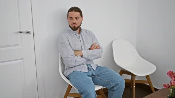 Nuori latino mies istuu tuolilla kädet ristissä ele ja vakava kasvot odotushuoneessa - Materiaali, video