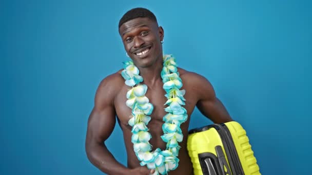 Afrikanischer amerikanischer Mann steht hemdlos in hawaiianischem Lei und hält Koffer über isoliertem blauem Hintergrund - Filmmaterial, Video