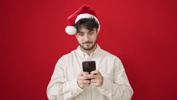 Νεαρός Ισπανός που φοράει χριστουγεννιάτικο καπέλο χρησιμοποιώντας smartphone σε απομονωμένο κόκκινο φόντο - Πλάνα, βίντεο