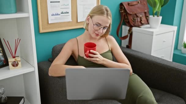 Νεαρή ξανθιά επιχειρηματίας χρησιμοποιώντας φορητό υπολογιστή πίνοντας καφέ στο γραφείο - Πλάνα, βίντεο