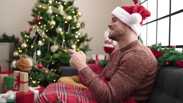 Молодой кавказский мужчина с помощью смартфона сидит на диване у рождественской елки дома - Кадры, видео