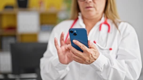 Lekarka w średnim wieku używająca smartfona z poważną twarzą w klinice - Materiał filmowy, wideo