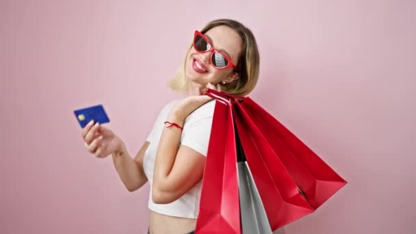 Jovem loira cliente segurando sacos de compras e cartão de crédito sobre fundo rosa isolado - Filmagem, Vídeo