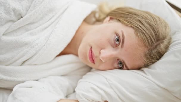 Młoda blondynka w szlafroku leżąca na łóżku uśmiechnięta do sypialni - Materiał filmowy, wideo