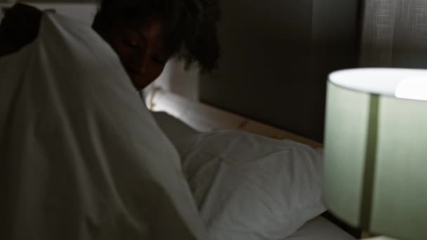 Afroamerikanerin liegt auf Bett und schaltet Lampe aus, um im Schlafzimmer zu schlafen - Filmmaterial, Video