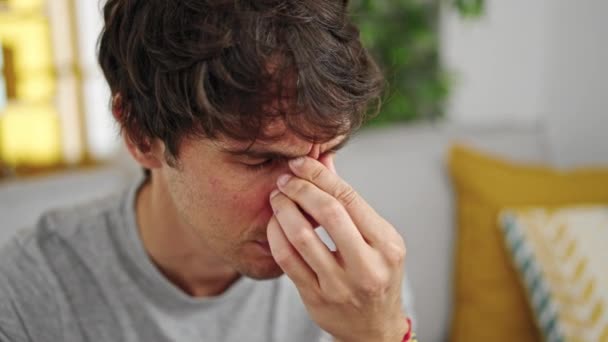 Молодий іспаномовний чоловік сидить на дивані потираючи ніс вдома - Кадри, відео