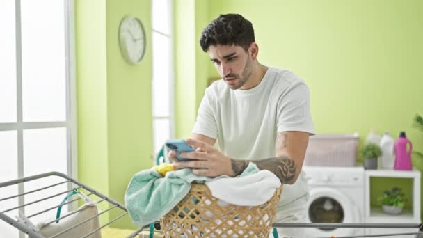 Молодий іспаномовний чоловік використовує смартфон, що спирається на кошик з одягом у пральні - Кадри, відео