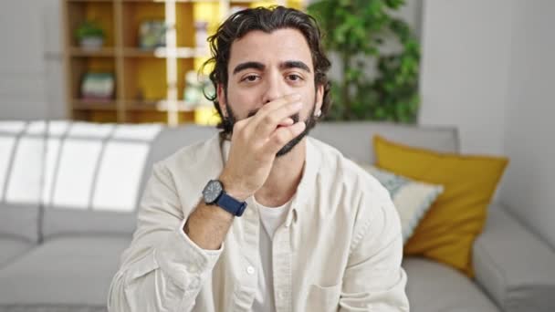 Молодий іспаномовний чоловік сидить на дивані робить німий жест вдома - Кадри, відео