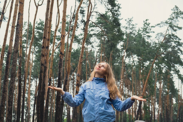 Happy young woman in blue raincoat enjoying the woods in park. True emotions open arms outdoors in rainy weather forecast. Tourist rest and feel freedom. Autumn season - Photo, image