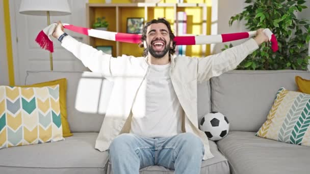 Joven hombre hispano apoyando equipo de fútbol sentado en sofá en casa - Metraje, vídeo