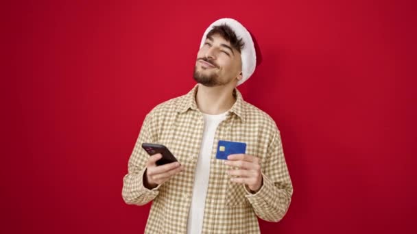 młody arabski człowiek robi Boże Narodzenie zakupy online ze smartfonem na izolowanym czerwonym tle - Materiał filmowy, wideo