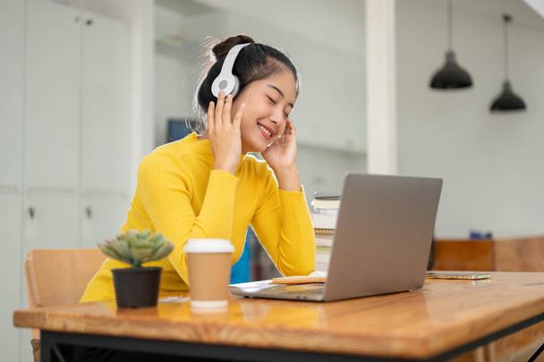 Μια όμορφη και χαρούμενη Ασιάτισσα φοιτήτρια απολαμβάνει να ακούει μουσική στα ακουστικά της ενώ κάνει τα μαθήματά της ή διαβάζει βιβλία σε μια βιβλιοθήκη. - Φωτογραφία, εικόνα