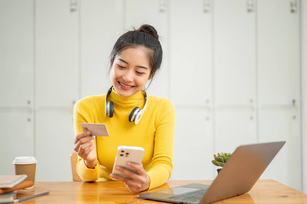 Una hermosa joven asiática sosteniendo una tarjeta de crédito o débito y su teléfono inteligente, utilizando una aplicación de banca móvil para transferir su dinero o pagar facturas en línea. - Foto, imagen