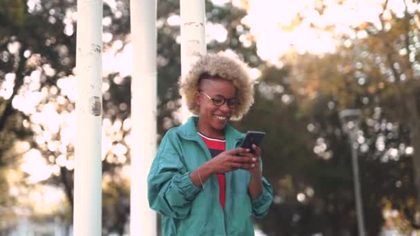 Pozitív fiatal afro-amerikai lány sportruházatban és szemüvegben mosolyog és nézi a képernyőt, miközben a városi parkban böngészi a mobiltelefont - Felvétel, videó