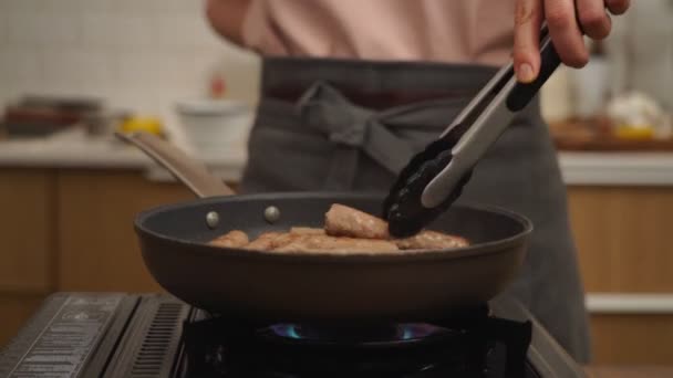Tempo reale di coltura anonima cuoco girando arrosto salsicce fatte in casa con pinze da cucina in padella volante posto sul fornello - Filmati, video