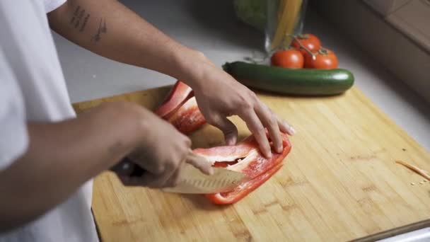 Masculino afroamericano irreconocible recortado en camiseta blanca rebanando pimiento rojo maduro con cuchillo afilado en tabla de cortar preparando ensalada - Metraje, vídeo