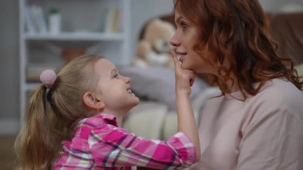 Positive woman talking as girl covering lips with finger and lady biting kid laughing out loud. Side view portrait of joyful carefree Caucasian mother and daughter having fun enjoying weekend at home - Séquence, vidéo