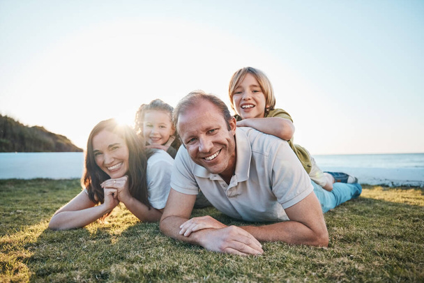 Family, parents and portrait of children on grass by ocean for bonding, relationship and relax together. Nature, sunlight and happy mother, father and kids on holiday, vacation and travel by sea. - Photo, Image