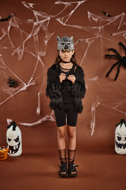 милая девушка в искусственном меховом одеянии с волчьей маской стоя на коричневом фоне, Хэллоуин концепции - Фото, изображение