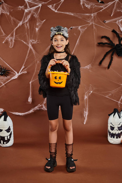 chica feliz sosteniendo cubo de dulces en máscara de lobo y traje negro, concepto de Halloween - Foto, imagen