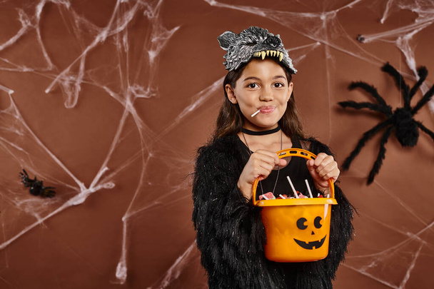 Nahaufnahme Mädchen mit Lutscher hält Süßigkeiteneimer auf braunem Hintergrund mit Spinnennetz, Halloween - Foto, Bild