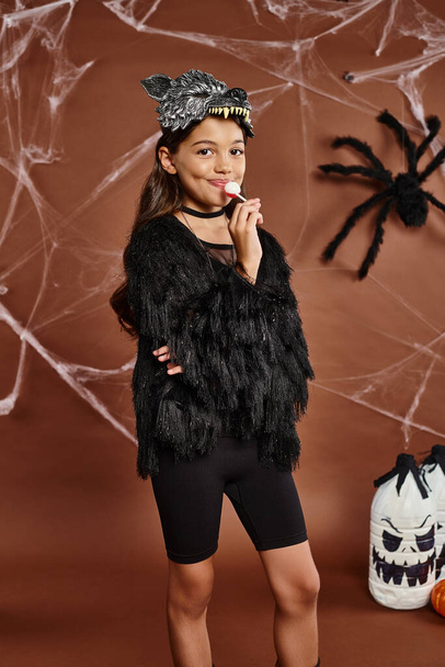 niña preadolescente con brazos cruzados y piruleta sobre fondo marrón con tela de araña, concepto de Halloween - Foto, imagen