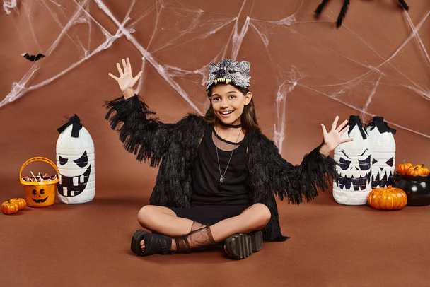 glückliches Mädchen in Wolfsmaske und schwarzem Gewand, sitzend mit überkreuzten Beinen und offenen Handflächen, Halloween - Foto, Bild