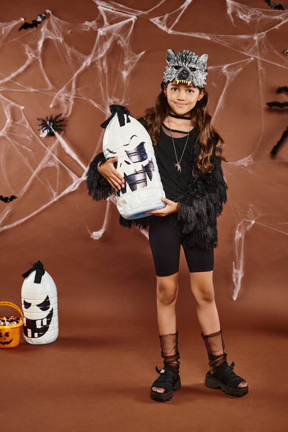smiley preteen girl holds Halloween lantern on brown backdrop with web, spiders and bats, Halloween - Photo, Image