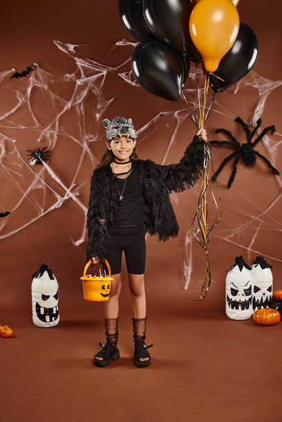 sonriente chica sostiene negro y naranja globos y cubo con dulces, concepto de Halloween - Foto, Imagen