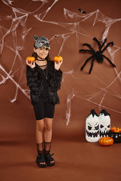 preteen girl standing in wolf mask holds pumpkins on brown backdrop, Halloween concept - Photo, Image