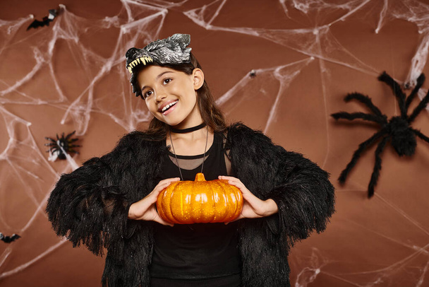 smiley preteen girl holding pumpkin in her hands on brown backdrop, Halloween concept - Foto, afbeelding