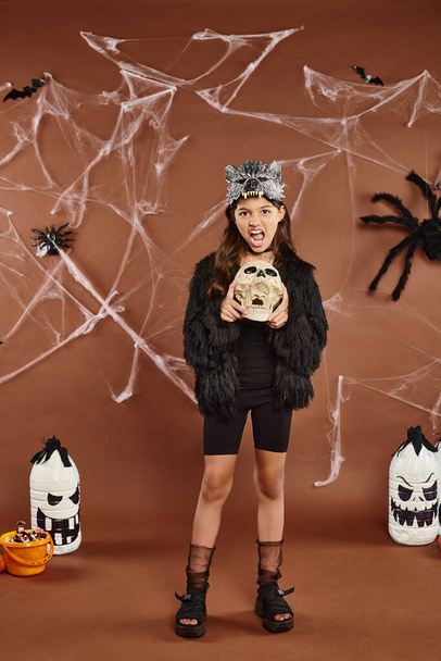 preteen girl grimacing and holding skull on brown backdrop with spiderweb and lanterns, Halloween - Photo, Image