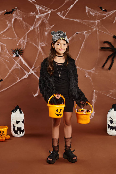 cheerful kid in wolf mask holds two buckets of sweets with bats and spiders on backdrop, Halloween - Photo, Image