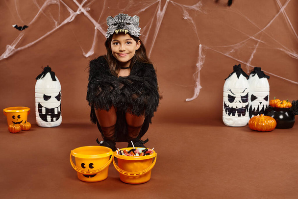 preteen kid squatting down near buckets of sweets with lanterns and pumpkins on backdrop, Halloween - Photo, Image